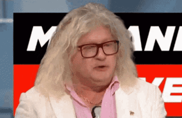 a man with long blonde hair and glasses stands in front of a sign with the word mani on it