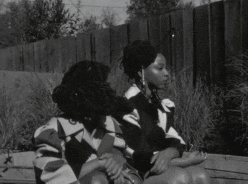 a black and white photo of two women sitting next to each other in front of a fence