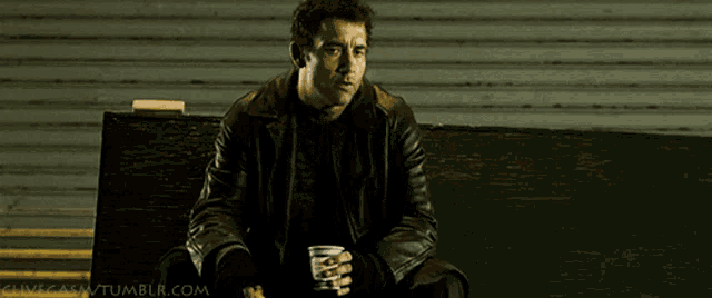 a man in a leather jacket sits on a bench with a cup of coffee in his hand