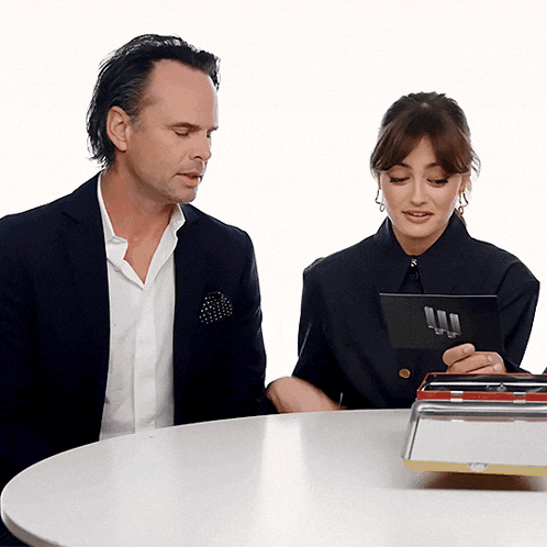a man and a woman sit at a table looking at a tablet with the letter l on it