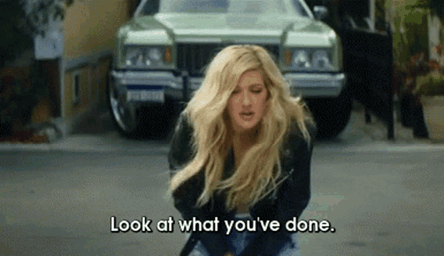 a blonde woman is standing in front of a green car and saying look at what you 've done .