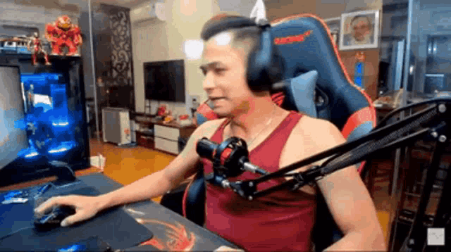 a man wearing headphones and a red tank top is sitting in front of a computer and a microphone .