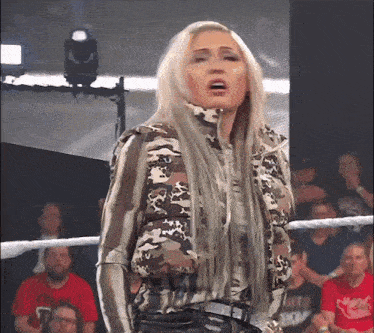 a woman wearing a camo jacket stands in a wrestling ring