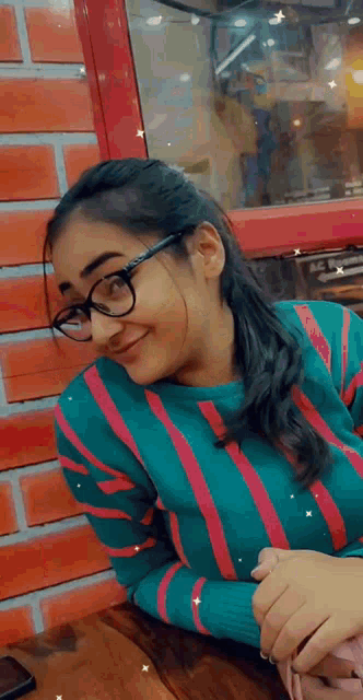 a woman wearing glasses and a striped sweater is sitting at a table in front of a brick wall .