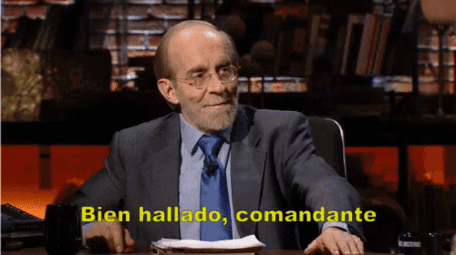 a man in a suit and tie is sitting at a table with the words bien hallado commandante written on the screen