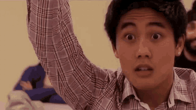 a young man in a plaid shirt is raising his hand in a classroom to answer a question .