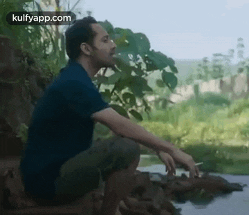 a man is smoking a cigarette while sitting on a rock next to a river .