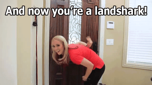 a woman is standing in front of a door with the words and now you 're a landshark written above her