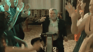a man in a suit is sitting in a wheelchair surrounded by people dancing .