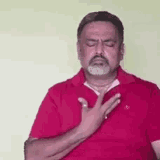 a man with a beard is wearing a red shirt and making a gesture with his hand on his chest .