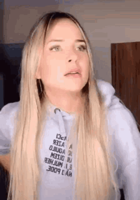 a woman with long blonde hair and green eyes is wearing a white hoodie and making a funny face .