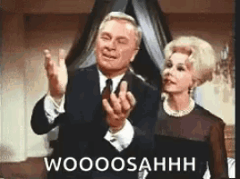 a man and woman are standing next to each other and the man is giving the middle finger while the woman looks on .