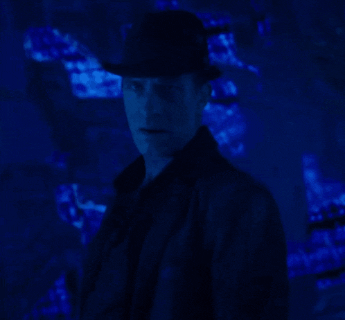 a man wearing a hat and a jacket stands in front of a blue background