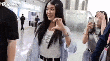 a girl is making a heart shape with her hands in an airport while a man takes a picture of her .