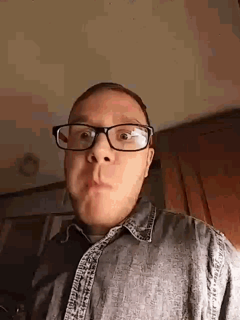 a man wearing glasses and a denim shirt is making a face .