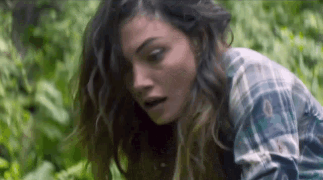 a woman in a plaid shirt is standing in the woods and looking down .