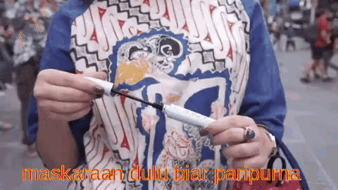 a woman wearing a batik shirt is holding a mascara in her hand