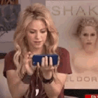 a woman is holding a cell phone in her hand in front of a poster of shakira .