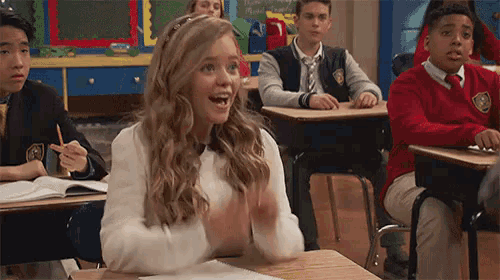 a girl is sitting at a desk in a classroom clapping her hands .