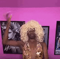 a woman in a wig is standing in front of a pink wall with her arms in the air .