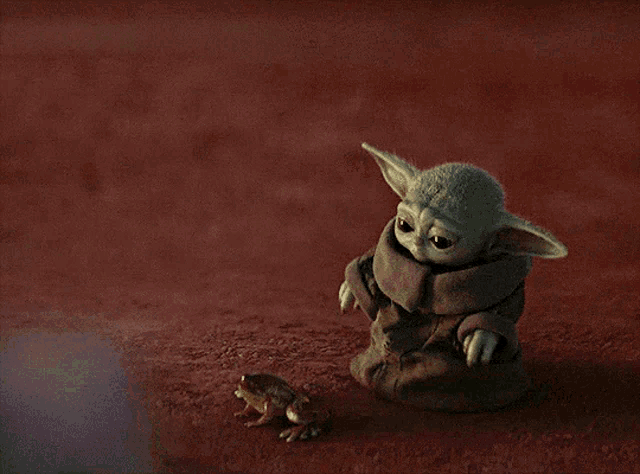 a baby yoda sits next to a small frog on a red surface