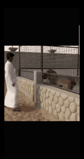 a man in a white robe is standing next to a fence with a dog in it .