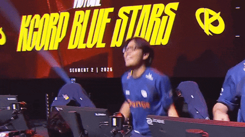a man wearing a blue shirt that says orange is dancing in front of a sign that says kcorp blue stars