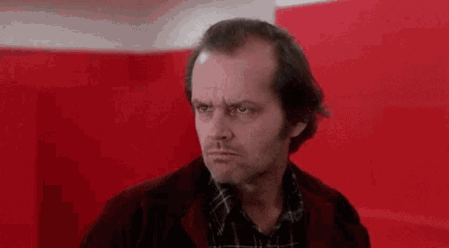 a man in a red jacket and plaid shirt is standing in front of a red wall and looking at the camera .