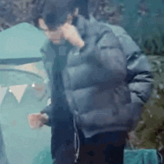 a man in a black jacket is talking on a cell phone while standing in front of a tent .