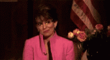a woman in a pink suit and glasses is smoking a cigarette in front of a flag .
