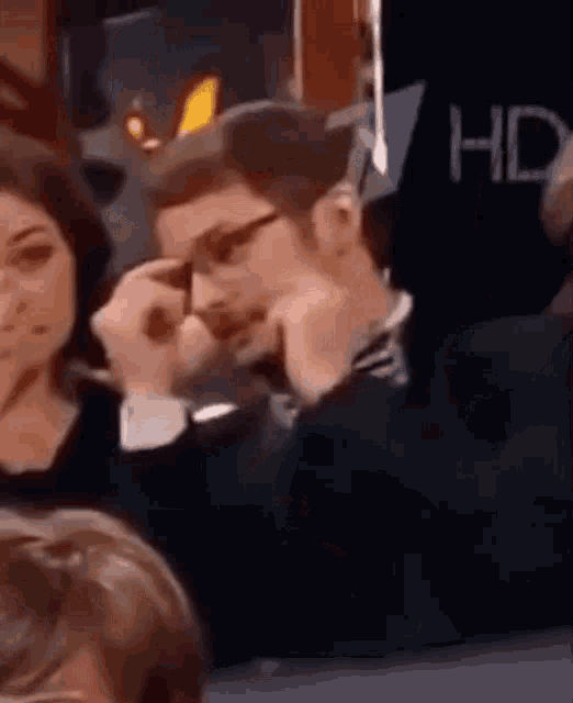 a man wearing glasses is sitting in a crowd of people with his hands on his chin .