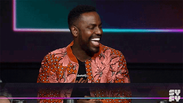a man in a floral jacket is laughing in front of a tv screen that says sy