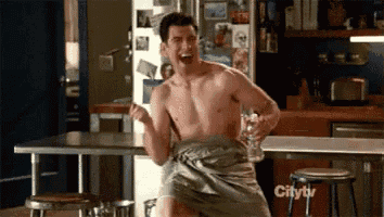 a shirtless man wrapped in a towel is holding a glass of beer in a kitchen .