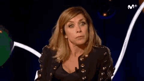 a woman in a black jacket with gold studs is sitting in front of a blue background and making a funny face .