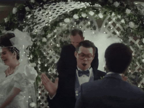 a man in a tuxedo and bow tie stands in front of a bride and groom at their wedding