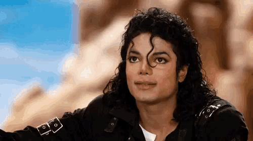 a close up of a man with curly hair wearing a black jacket and looking at the camera .