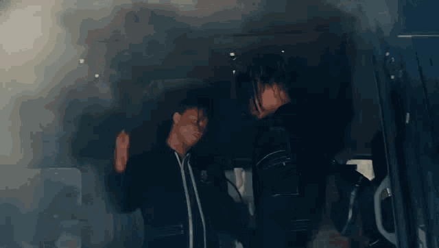 two men are standing in the back of a car smoking a cigarette .