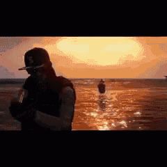 a man in a baseball cap is standing in the water at sunset