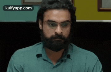 a man with a beard and glasses is wearing a green shirt and looking at the camera .
