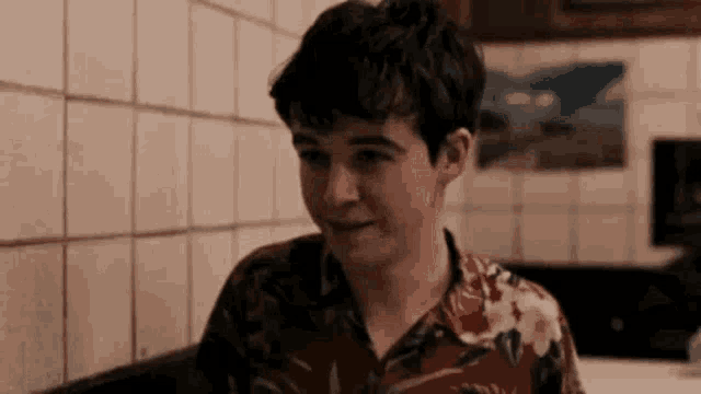 a young man in a floral shirt is sitting at a table in a kitchen .