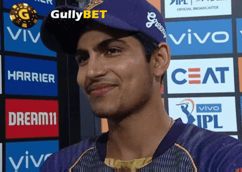 a man wearing a purple hat stands in front of a wall of advertisements including ceat and dream11