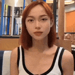 a woman with red hair is wearing a white tank top and looking at the camera .
