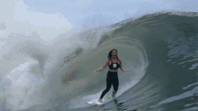a woman is riding a wave on a surfboard while wearing a black top with the letter r on it