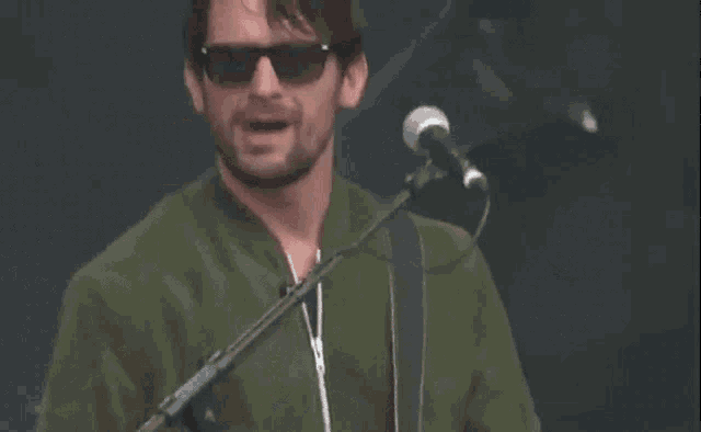 a man wearing sunglasses and a green jacket sings into a microphone