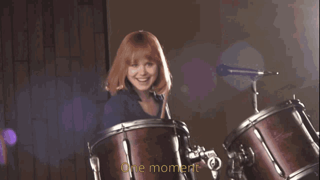 a woman playing drums with the words " one moment " written below her