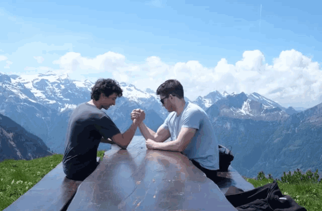 two men arm wrestling on a picnic table in front of mountains