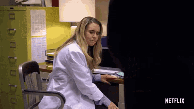 a woman in a lab coat is sitting at a desk with netflix written on the bottom