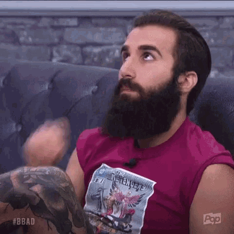 a man with a beard and a tattoo on his arm is sitting on a couch wearing a pink shirt .