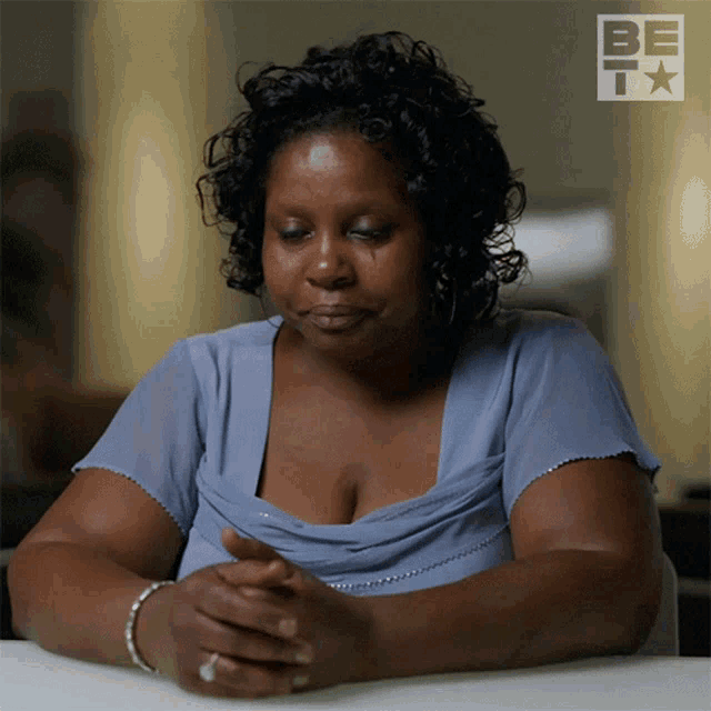 a woman in a blue dress is sitting at a table with her hands folded in front of a be tv logo