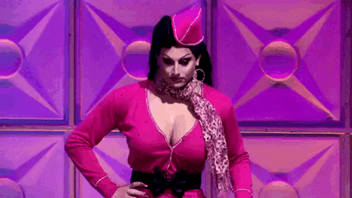 a drag queen in a pink dress and hat is standing in front of a purple wall .